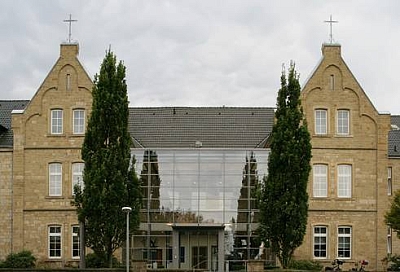 Das Bild zeigt den verglasten Haupteingang der Rehaklinik. Das Mauerwerk ist aus Sandstein. Neben dem Eingang stehen zwei große Bäume. Das Bild zeigt den verglasten Haupteingang der Rehaklinik. Das Mauerwerk ist aus Sandstein. Neben dem Eingang stehen zwei große Bäume.