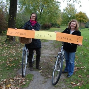 Zwei Damen mit Fahrrädern halten ein Schild hoch mit der Aufschrift: gemeinsam aktiv werden!