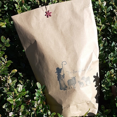 Eine braune gefüllte Tüte liegt in grünem Buchsbaum. Auf der Tüte sind ein Hirt und Schafe aufgedruckt. Eine braune gefüllte Tüte liegt in grünem Buchsbaum. Auf der Tüte sind ein Hirt und Schafe aufgedruckt.
