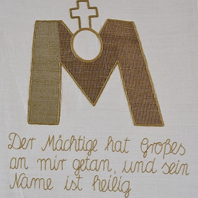 Gesticktes Bild im Hungertuch: Aus dem goldenen „M“ für Maria bricht die runde Hostie hervor, auf der das Kreuz steht.