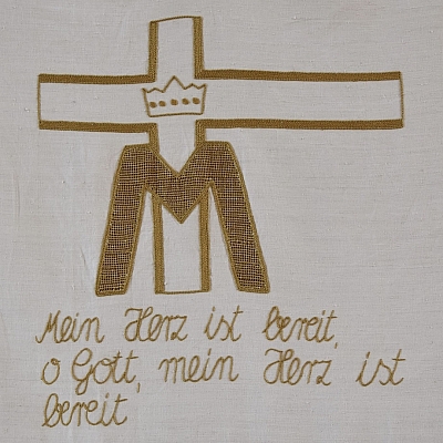 Gesticktes Bild im Hungertuch: Ein großes M für Maria und darüber eine Krone. Dahinter das Kreuz.