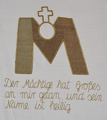 Gesticktes Bild im Hungertuch: Aus dem goldenen „M“ für Maria bricht die runde Hostie hervor, auf der das Kreuz steht.