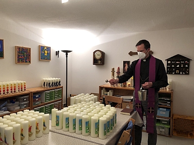 Eine große Menge fertig verzierter Kerzen stehen sortiert auf dem Tisch und dem Regal. Pastor Holtmann segnet die Kerzen.