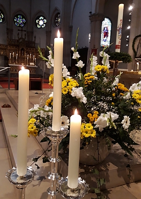 Drei brennende Altarkerzen stehen vor einem Blumengesteck in der St.-Agatha-Kirche